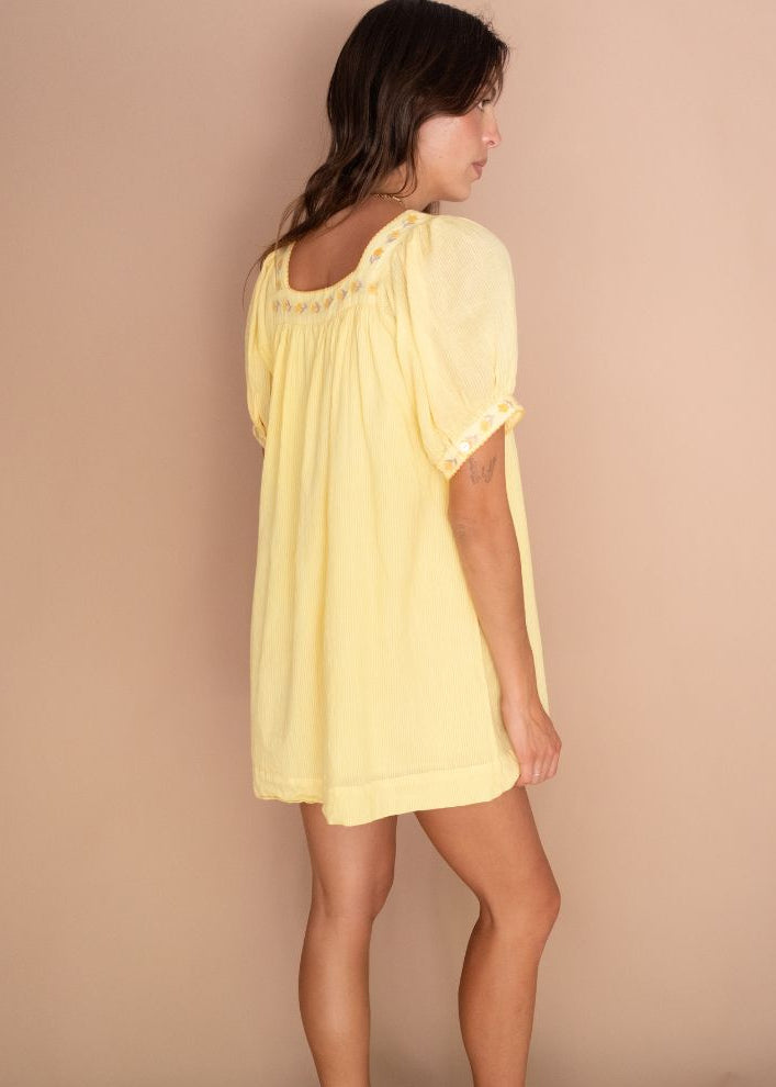 A woman wearing a pale yellow short-sleeve tunic dress with lace trim, shown from a side-back angle against a solid beige studio backdrop.
