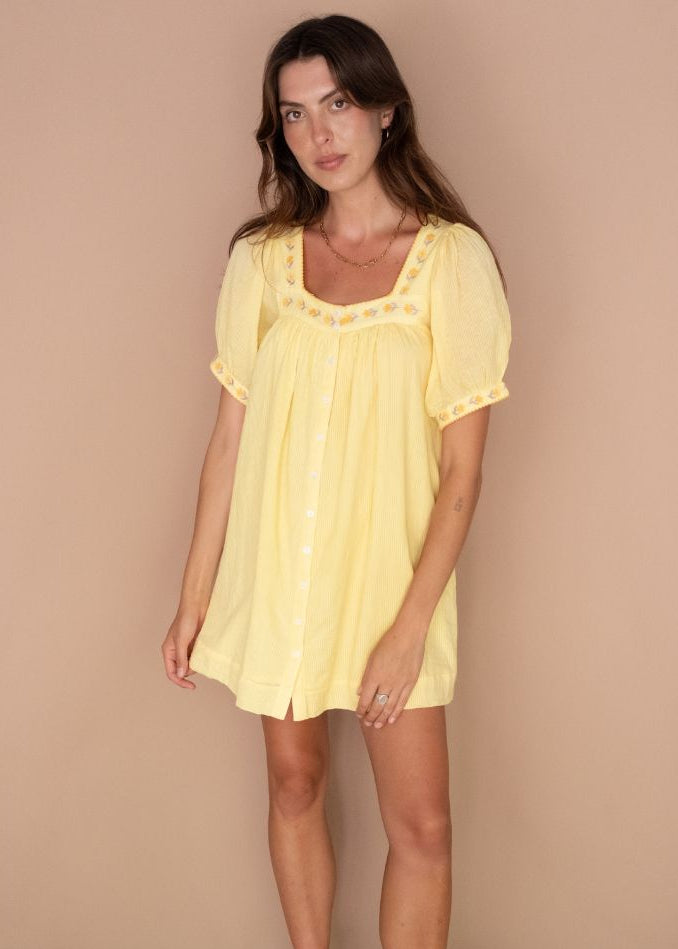 A woman modeling a pale yellow short-sleeve babydoll-style dress with a square neckline, decorative trim, and front buttons, standing against a plain beige studio backdrop.