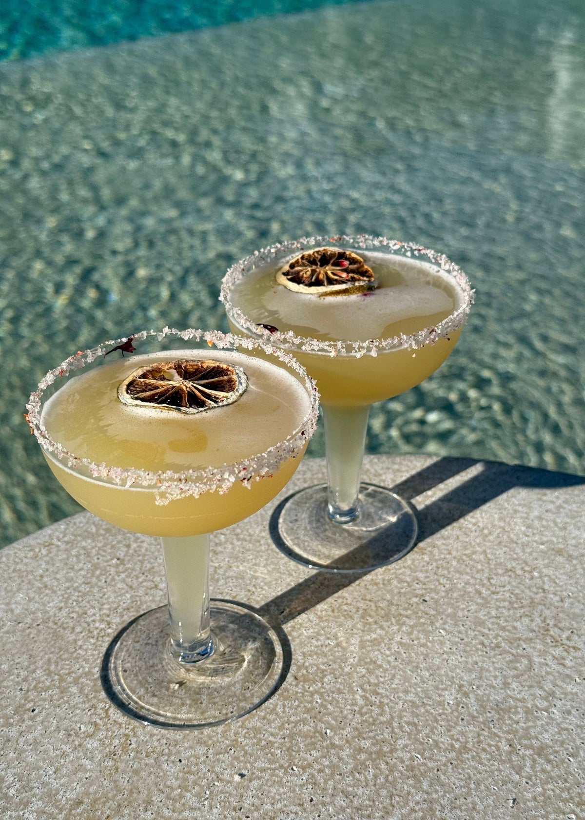 Two margarita cocktails with salt rims and lime wedges on a reflective pool surface.