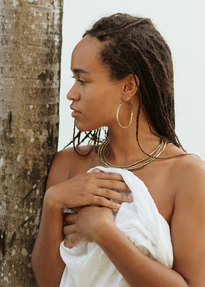 Woman with braided hair wearing gold hoop earrings and necklaces against a white wall.