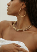 Woman wearing gold hoop earrings and necklaces against a neutral background
