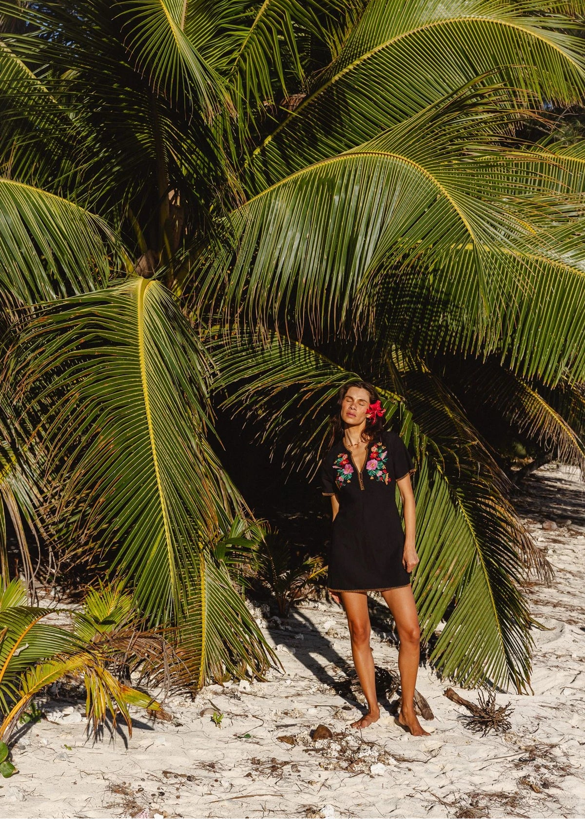 A woman stands on white sand beneath large green palm trees, wearing a black short-sleeve dress with colorful floral embroidery on the chest.