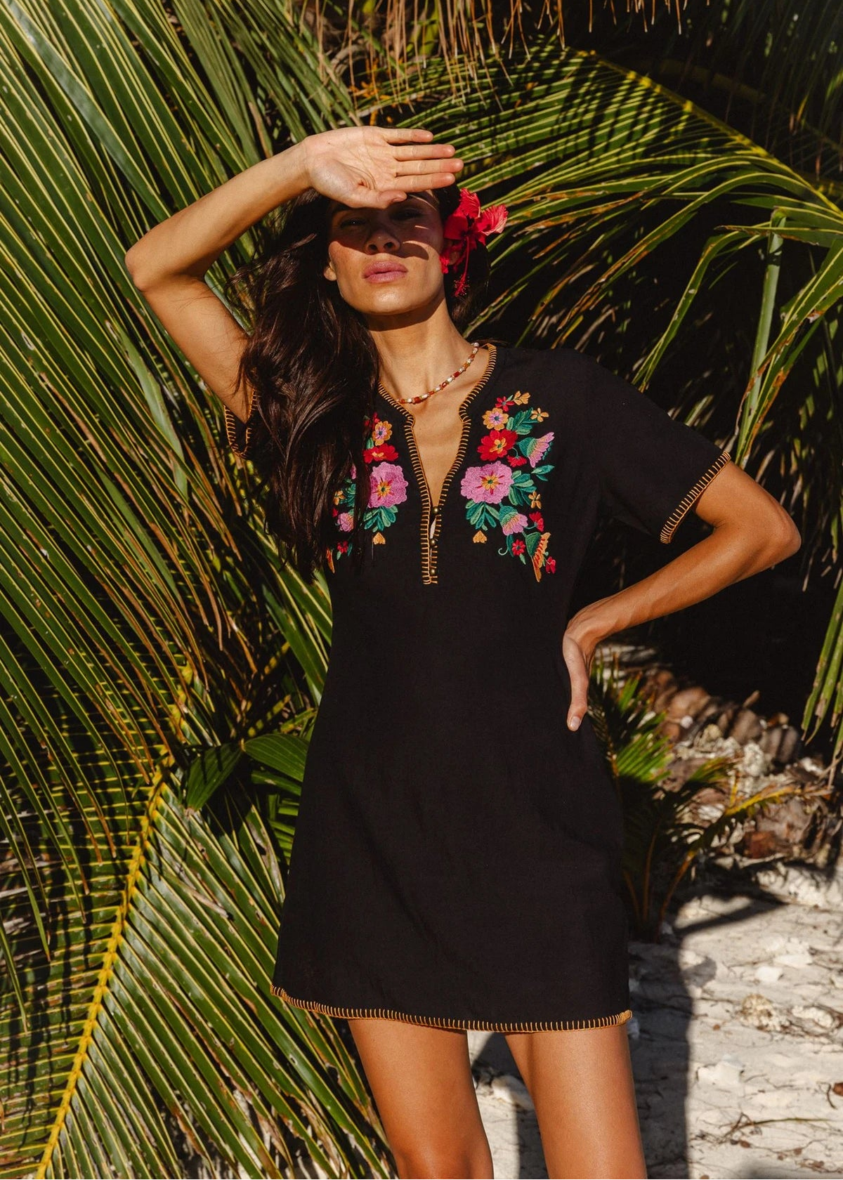 A woman wearing a black short-sleeve mini dress with colorful floral embroidery on the chest stands outdoors in front of large green palm leaves, with one hand shielding her eyes.