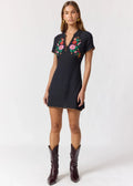 A woman modeling a short-sleeve black denim dress with colorful floral embroidery on the chest, paired with dark brown cowboy boots, against a plain white studio background.