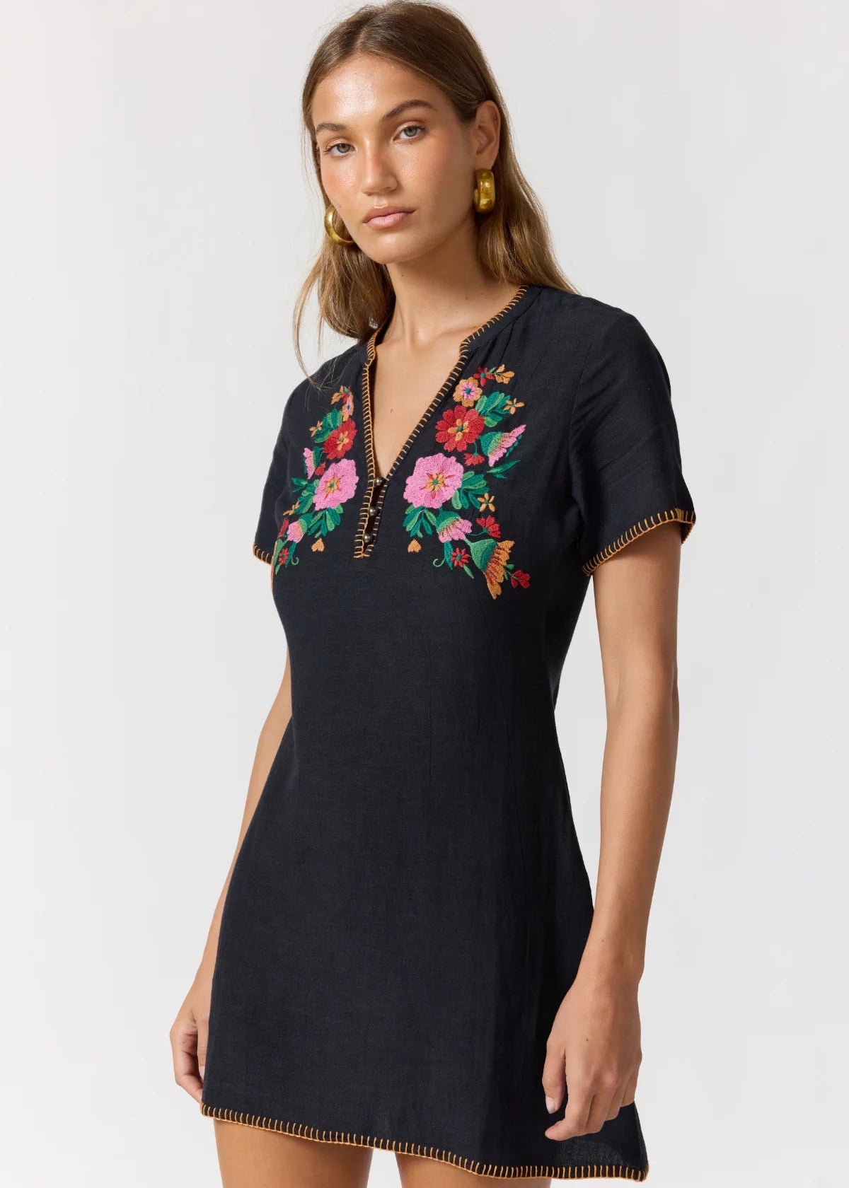 A woman modeling a black short-sleeve tunic dress with colorful floral embroidery around the V-neckline, photographed against a plain light gray studio background.
