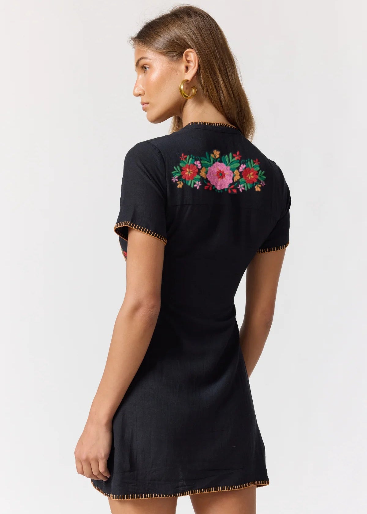 A woman shown from the back wearing a black short-sleeve dress with colorful floral embroidery across the upper back and gold-tone trim along the sleeve cuffs and hem.