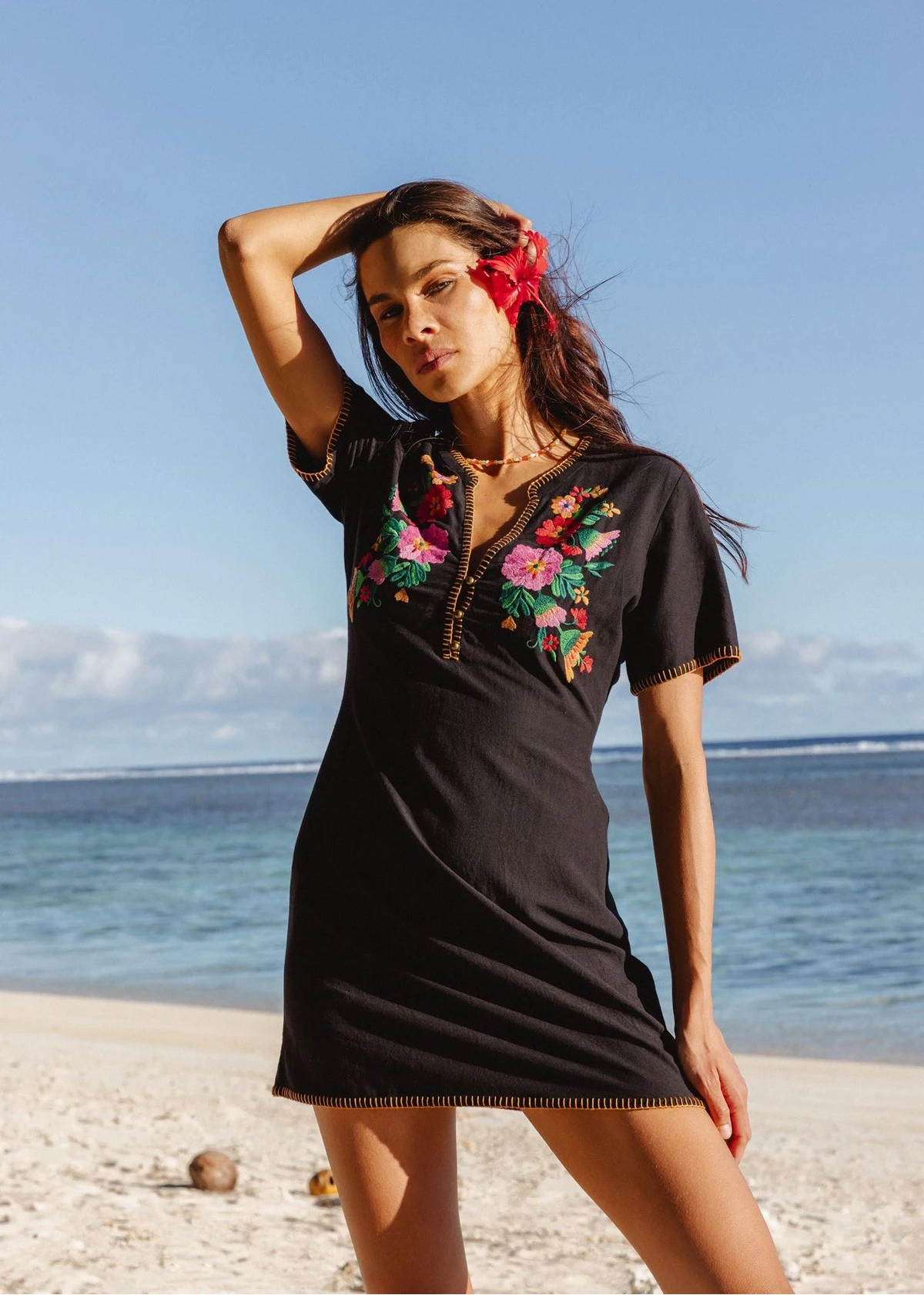 Woman in a black dress with floral embroidery standing on a beach.