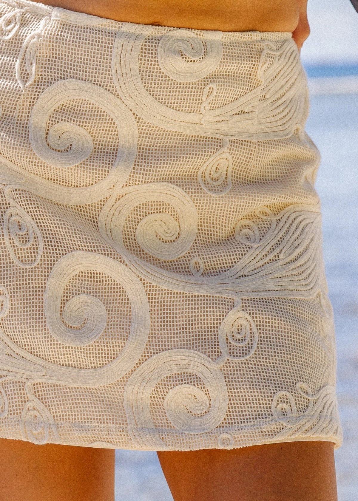 A close-up of a person wearing a white lace mini skirt with a textured mesh base and raised swirl patterns, photographed outdoors with a blurred beach background.