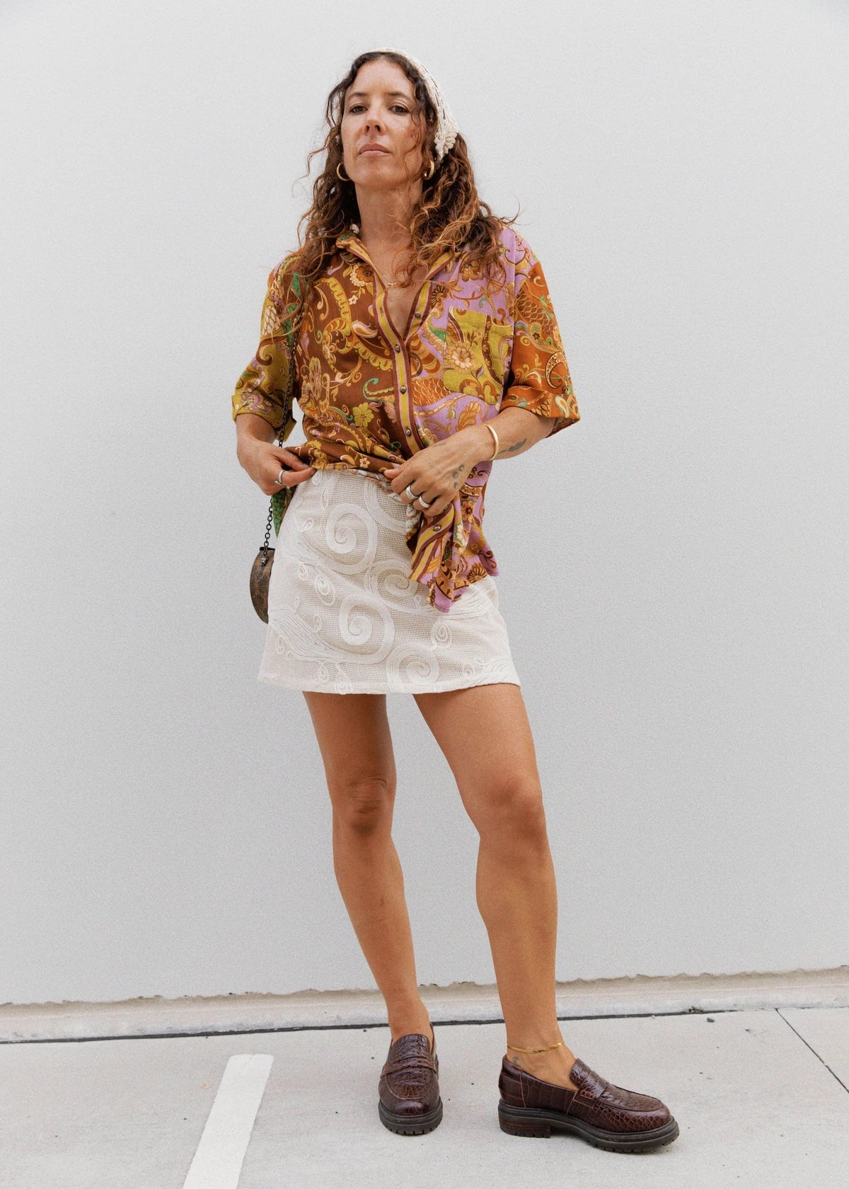 A woman stands against a plain light gray wall wearing a multicolor patterned button-up shirt tied at the waist, a white mini skirt, and dark brown leather loafers.