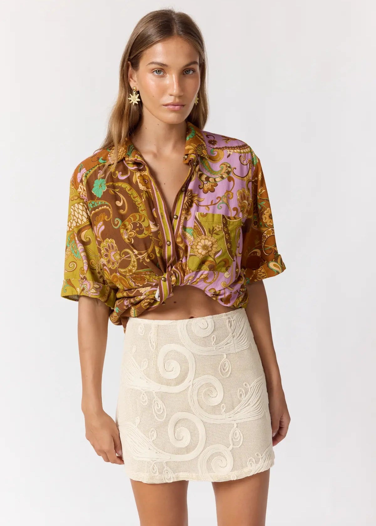 A model wearing a brown and purple paisley patterned short-sleeve button-up shirt tied at the waist, paired with a cream-colored lace mini skirt, against a plain white studio background.