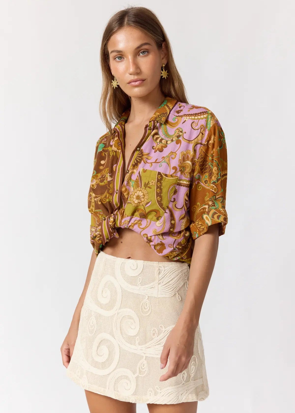 A model wearing a short-sleeve cropped button-up shirt with a brown and pink baroque pattern, tied at the waist, paired with a cream-colored embroidered mini skirt.