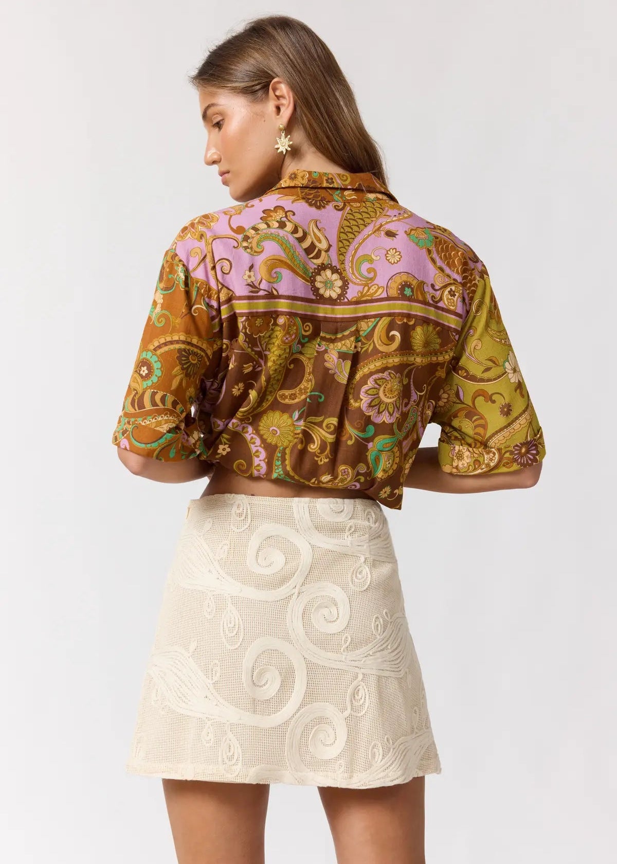 A woman shown from the back wearing a short-sleeve collared shirt with a colorful paisley print in brown, gold, and lavender, paired with a cream embroidered mini skirt, against a light gray studio background.