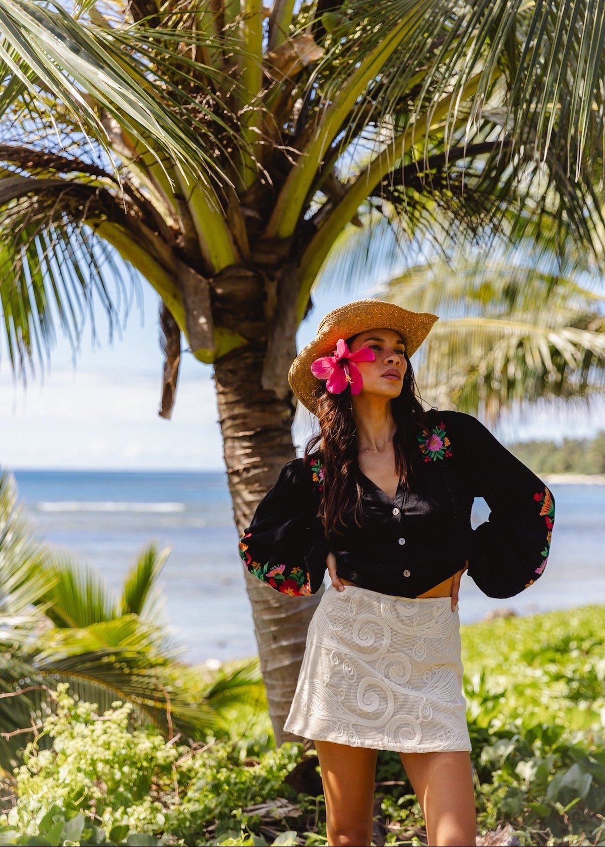 A woman posing outdoors on a tropical beach with palm trees and ocean in the background, wearing a black embroidered long-sleeve crop top, a white patterned mini skirt, and a straw hat with a pink flower.