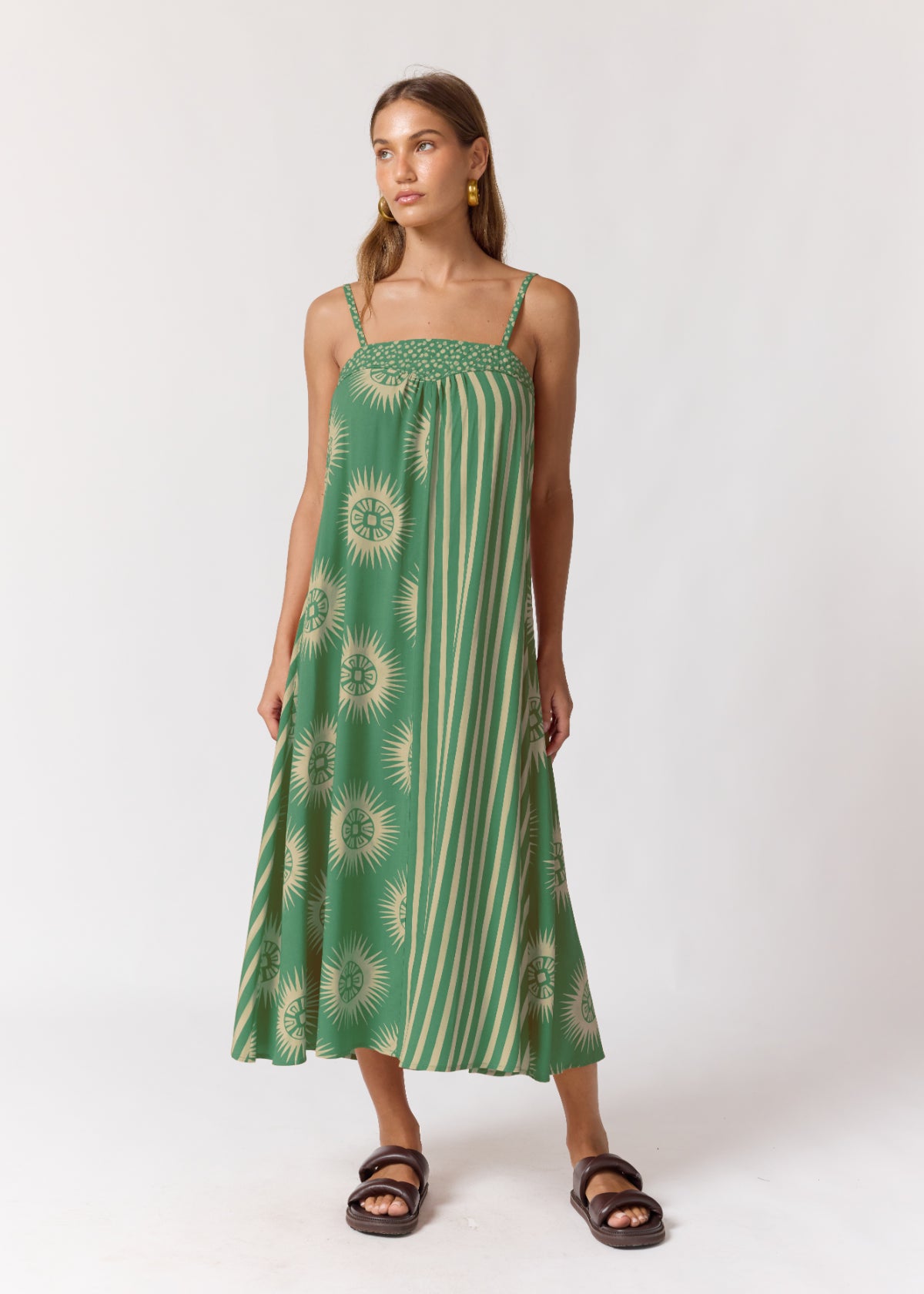 A woman modeling a green midi dress with a smocked bodice, featuring vertical stripes and sunburst floral patterns, paired with dark brown slide sandals against a plain white studio background.