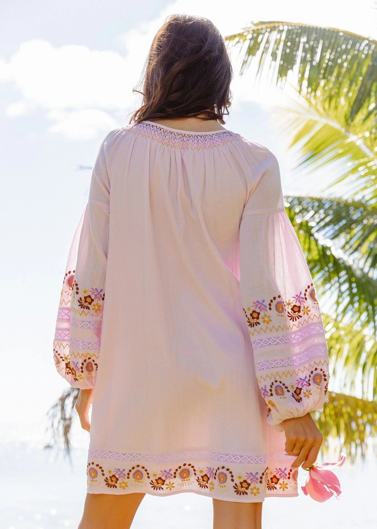 A woman viewed from behind wearing a light pink bohemian-style tunic dress with embroidered floral details on the sleeves and hem, set against a tropical beach background with palm trees.