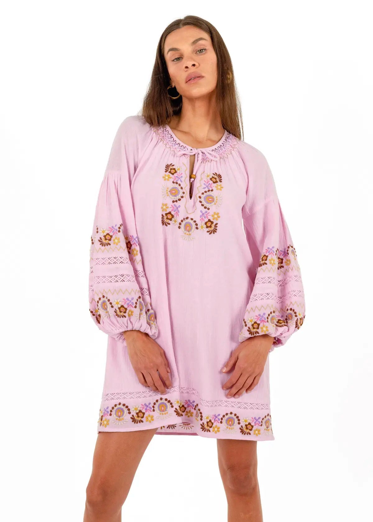 A woman modeling a light pink bohemian-style tunic dress with intricate multicolor floral embroidery on the neckline, sleeves, and hem, set against a plain white background.