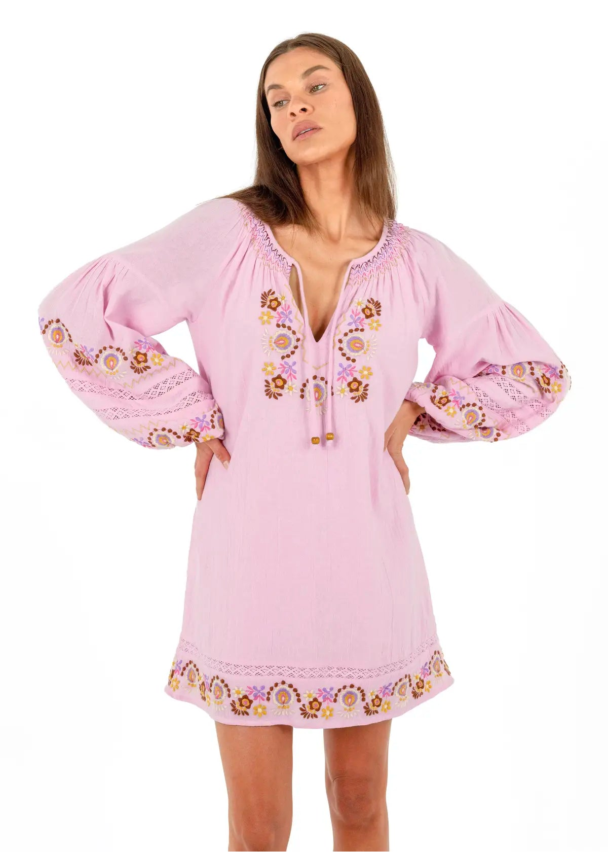 A woman modeling a light pink bohemian-style tunic dress with voluminous sleeves, a drawstring tie neckline, and colorful floral embroidery on the chest and cuffs, set against a plain white background.