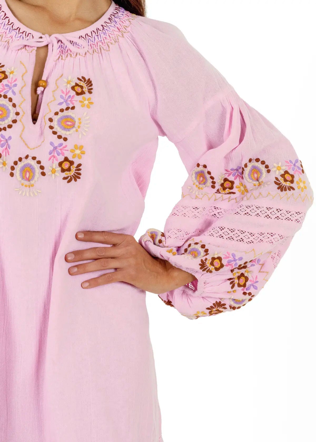 A close-up of a light pink long-sleeve tunic or dress with colorful floral embroidery and lace trim, worn by a model whose hand rests on the hip.