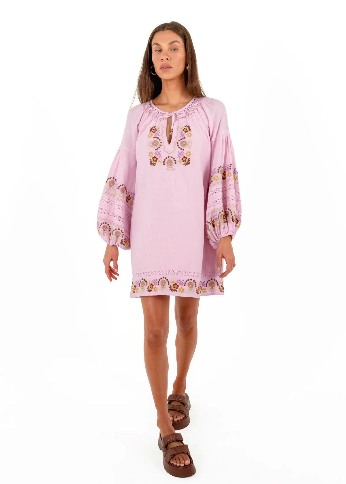 A woman modeling a light pink bohemian-style tunic dress with embroidered details on the chest and sleeves, paired with brown slide sandals, against a plain white background.