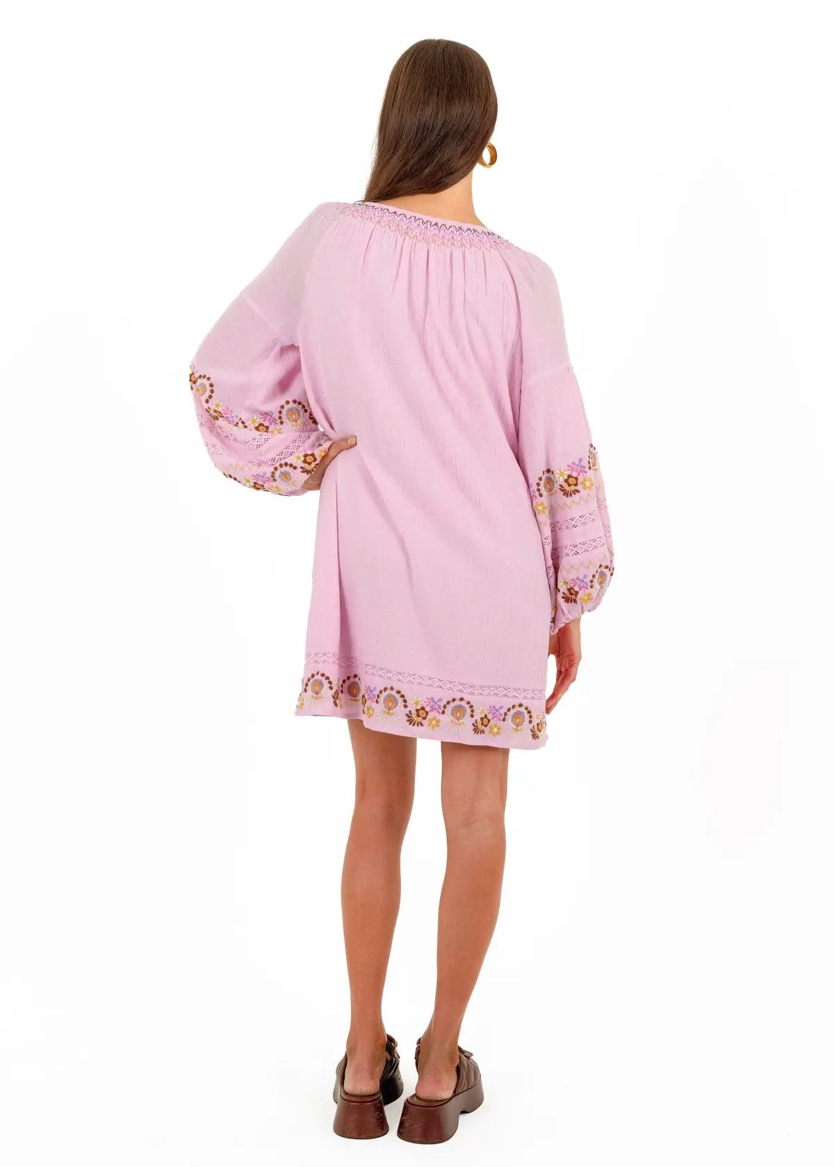 A woman viewed from the back wearing a light pink short dress with embroidered details on the sleeves and hem, paired with brown shoes, against a plain white background.