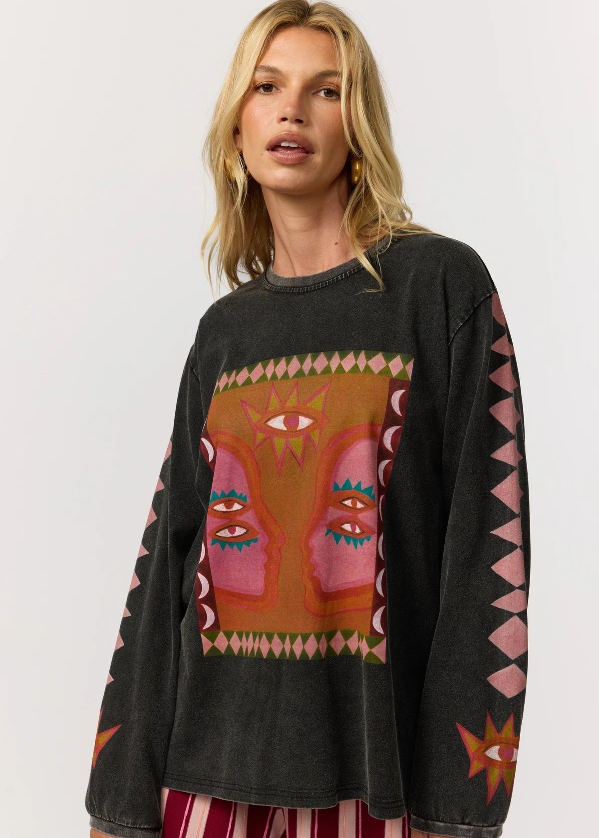 A woman wearing a black washed long-sleeve sweatshirt with a colorful Southwestern-style eye mask graphic on the front and pink geometric patterns on the sleeves, paired with red and white striped pants, against a light gray background.