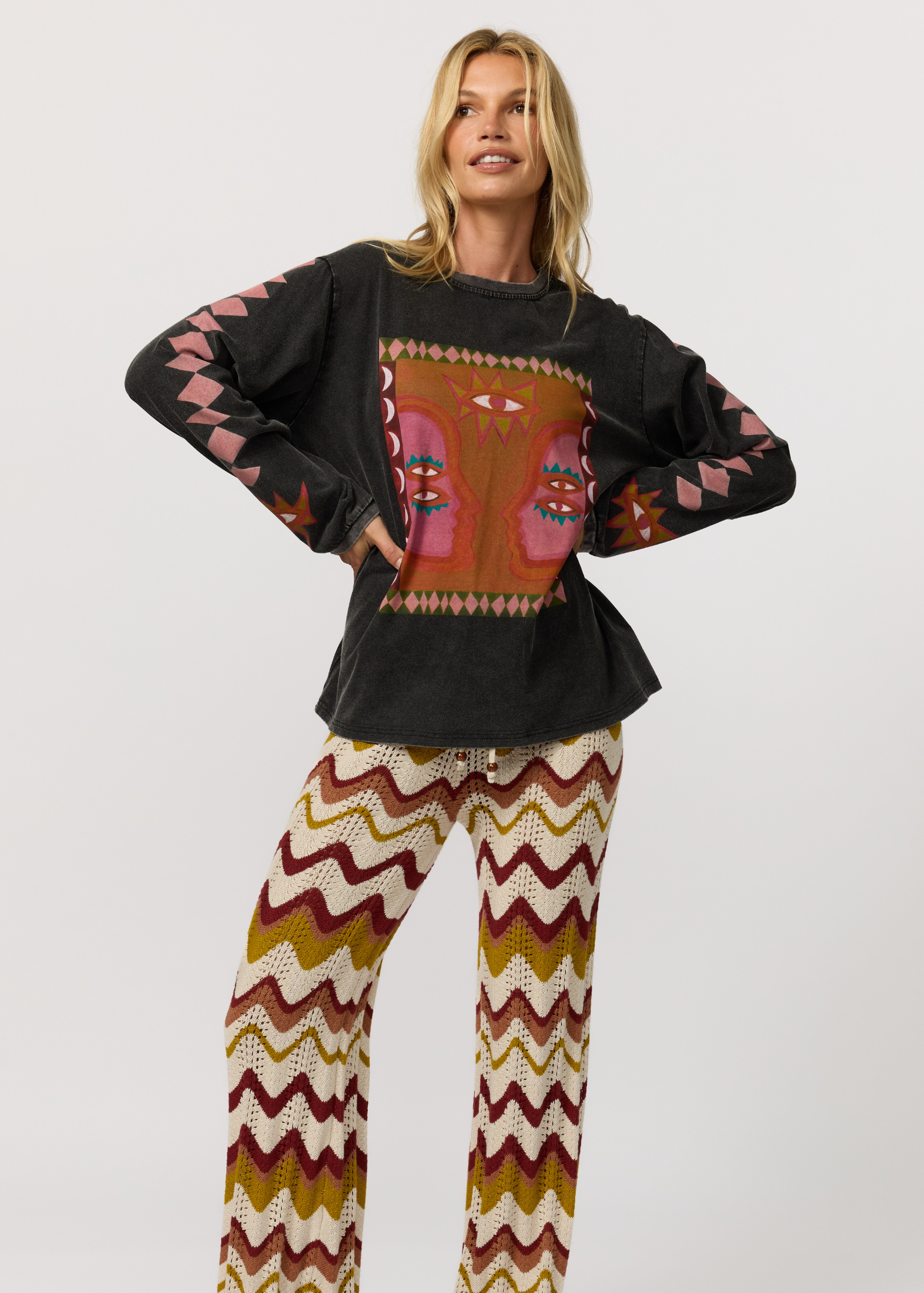A woman modeling a black long-sleeve top with a colorful geometric chest graphic and pink patterned sleeves, paired with matching zigzag-print pants in cream, rust, and burgundy, against a light gray studio background.