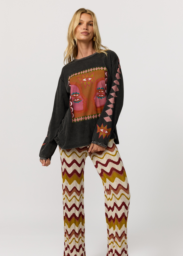 A woman modeling a black long-sleeve top with a colorful geometric and tribal-style graphic print on the front and sleeves, paired with patterned knit pants featuring a zigzag chevron design in cream, brown, and burgundy.