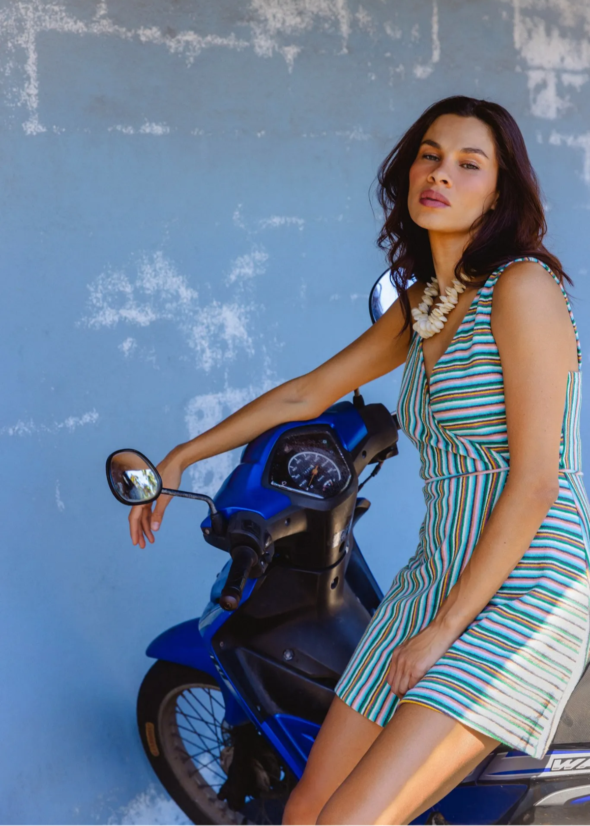 A woman wearing a green, white, and blue striped sleeveless dress sits on a blue scooter against a textured light blue wall.
