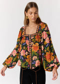 A woman wearing a long-sleeve floral blouse with a square neckline and colorful embroidered trim, paired with black pants, against a plain light gray studio background.