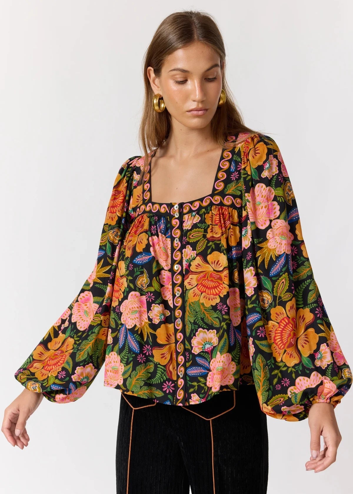 A woman wearing a long-sleeve floral blouse with a square neckline and colorful embroidered trim, paired with black pants, against a plain light gray studio background.