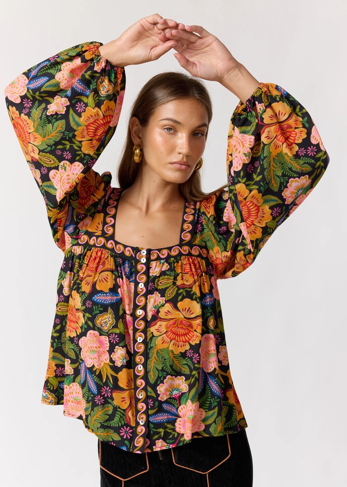A woman modeling a vibrant bohemian floral print blouse with dramatic bell sleeves, featuring orange, pink, yellow, and green flowers on a dark background.