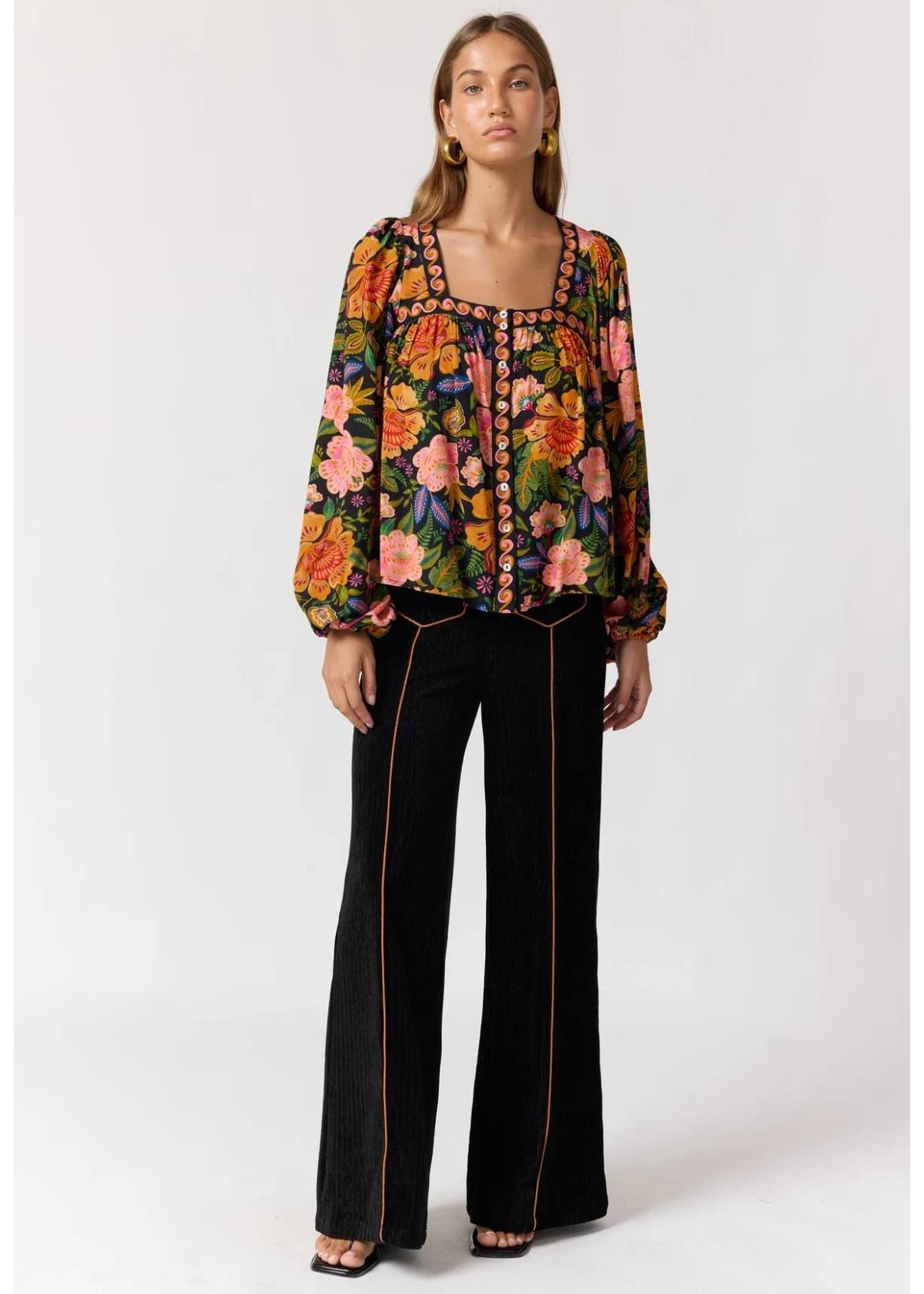 A woman modeling a long-sleeve floral blouse with a square neckline and black wide-leg pants with orange piping, standing against a light gray studio background.