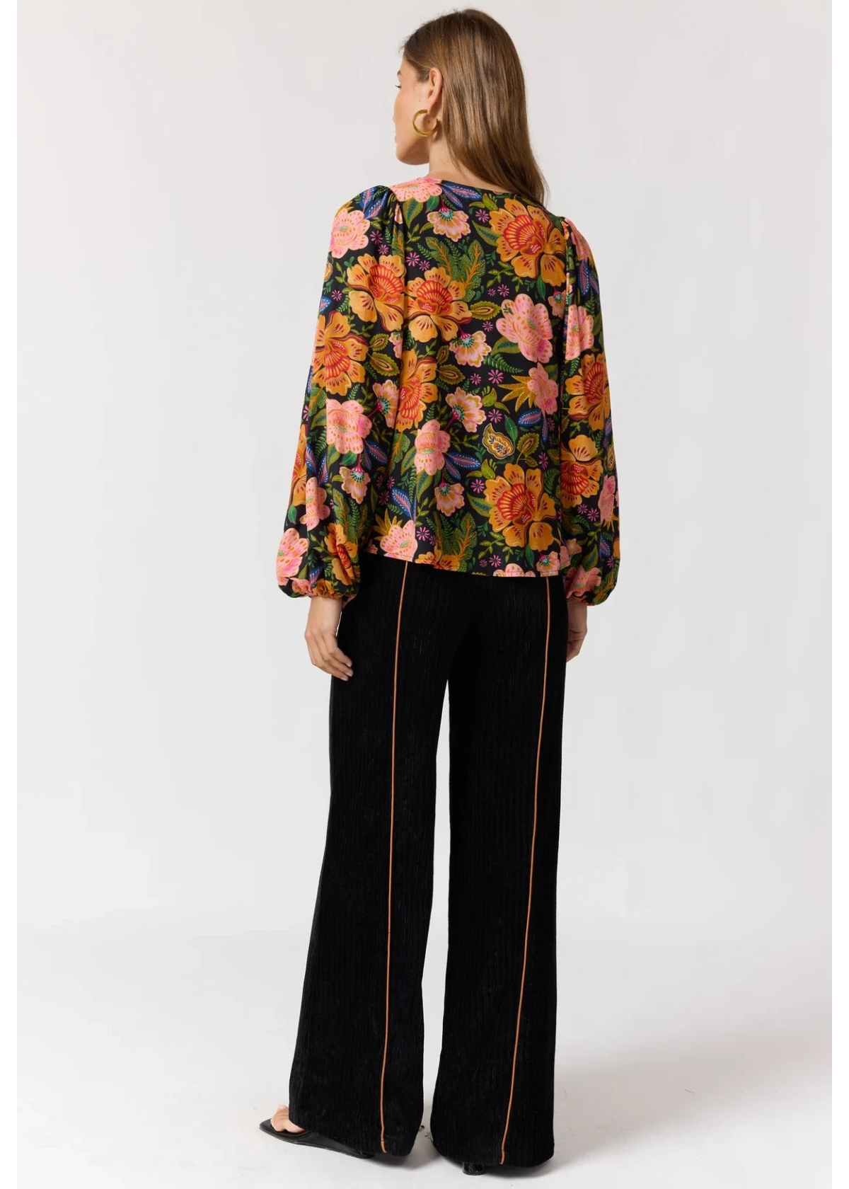 A woman shown from the back wearing a long-sleeve floral blouse with a black base and colorful orange, pink, and yellow flowers, paired with black wide-leg trousers with thin gold side piping, against a light gray studio background.