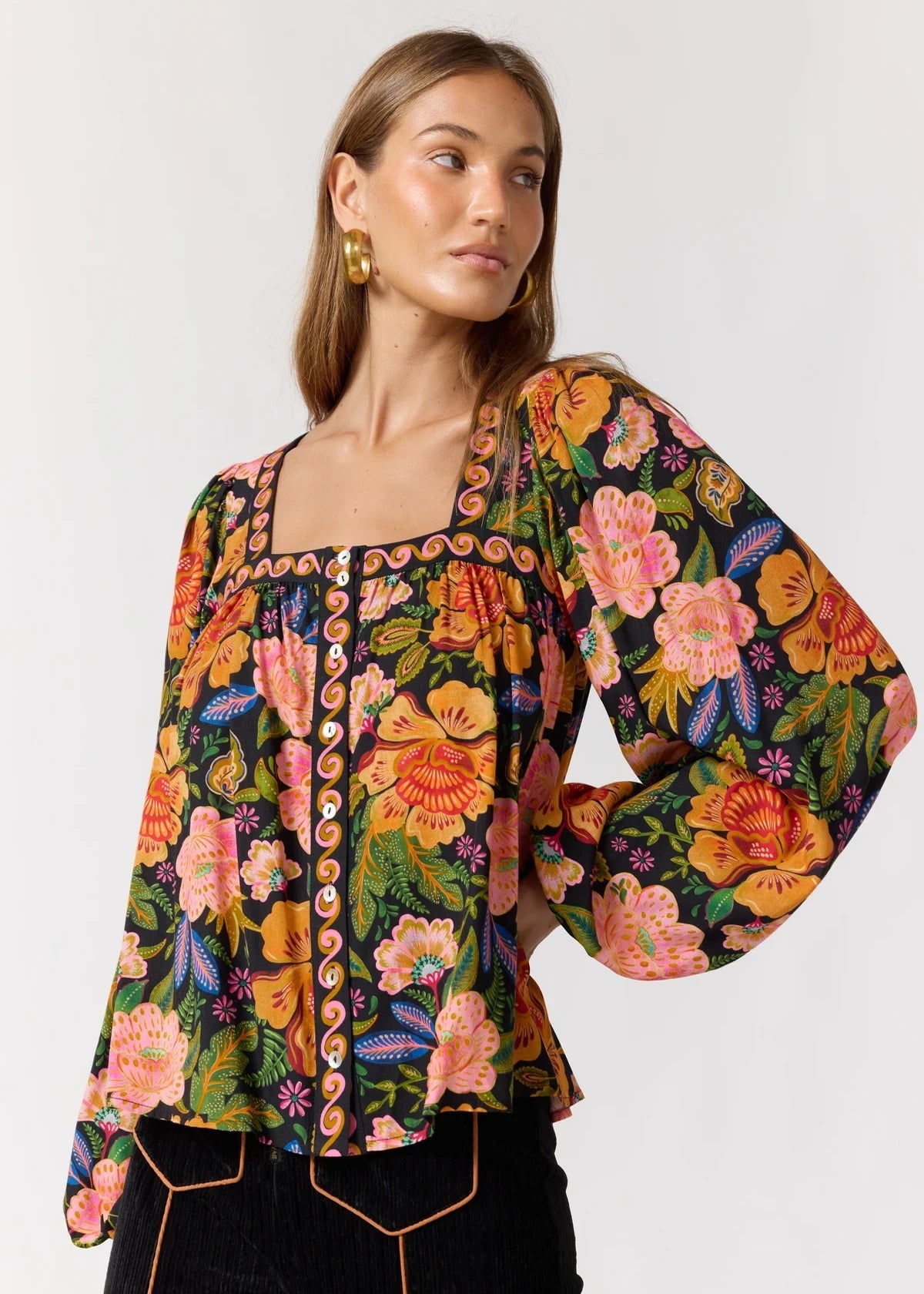 A woman wearing a vibrant, multicolor floral long-sleeve blouse with a square neckline and decorative trim, paired with black pants against a plain light gray background.