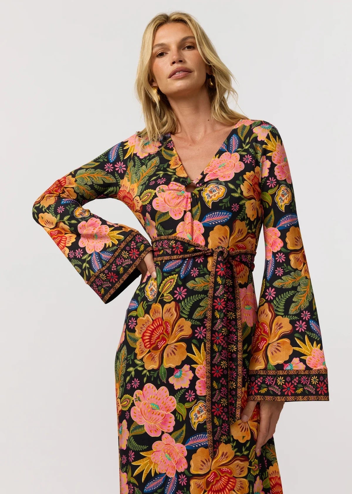 A woman modeling a long-sleeve wrap-style maxi dress with a vibrant multicolor floral print on a dark background, featuring a self-tie belt at the waist.