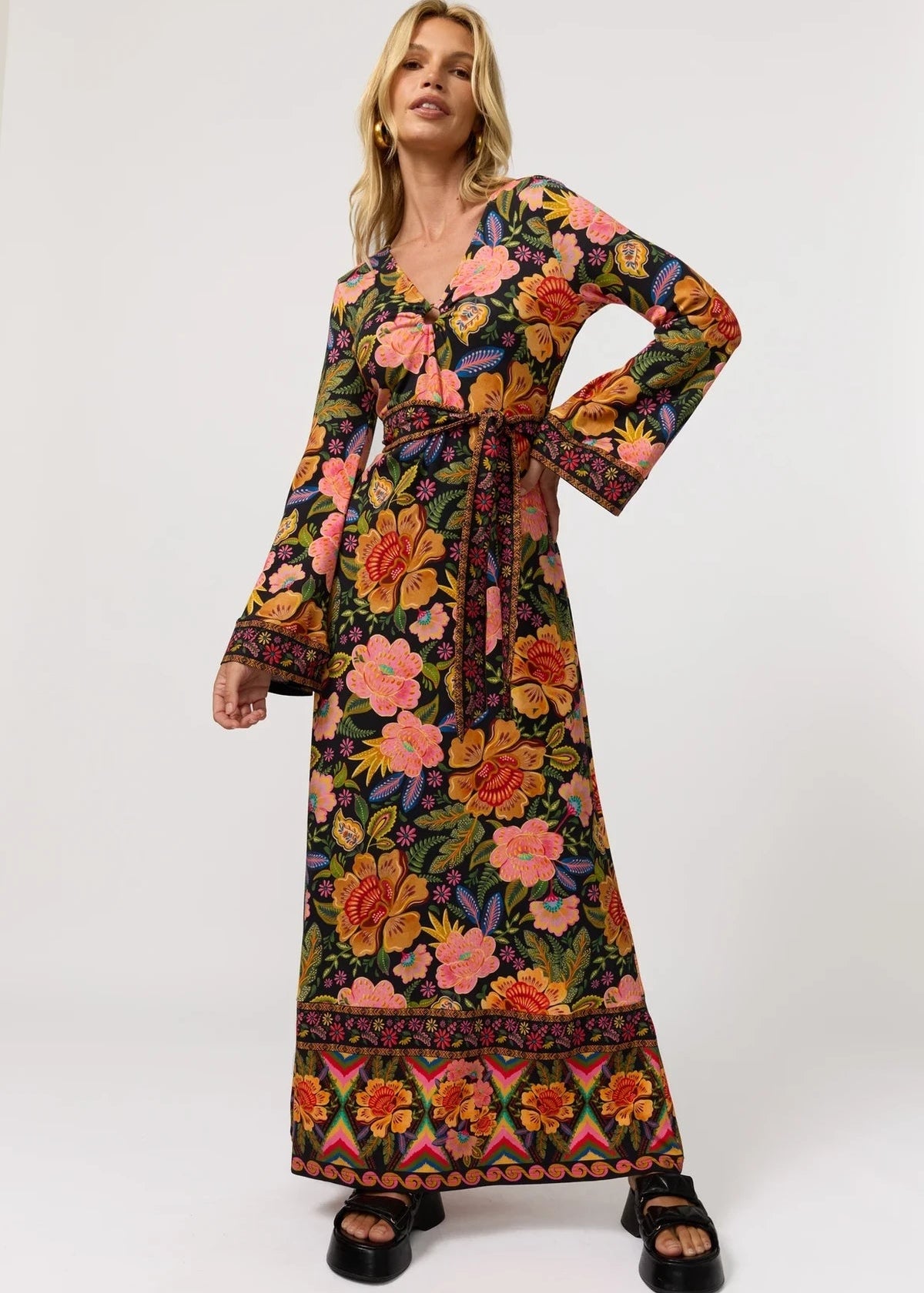 A woman modeling a long-sleeved maxi dress with a vibrant multicolor floral and geometric print on a dark background, paired with black platform sandals, against a plain white studio background.