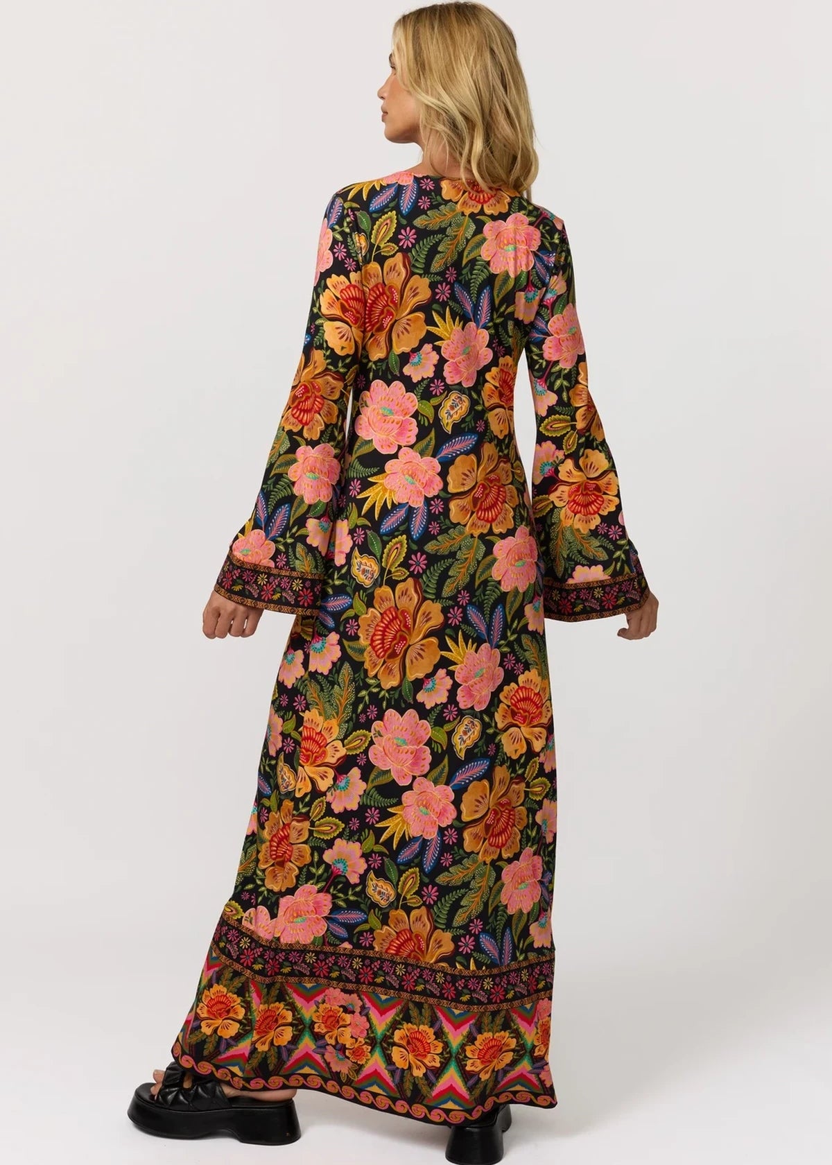 A woman shown from the back wearing a long-sleeved maxi dress with a vibrant multicolor floral pattern on a black background, paired with black shoes, against a plain light gray studio background.