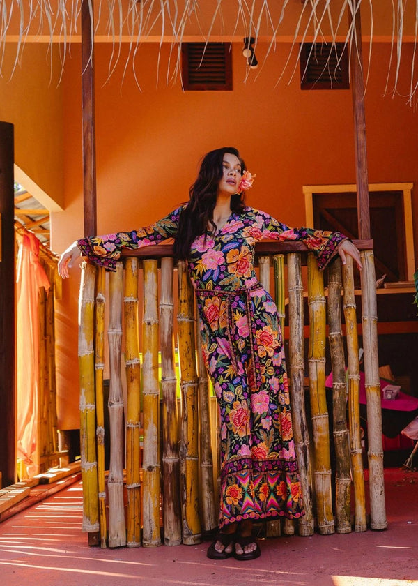 A woman wearing a colorful floral long-sleeve dress leans against a bamboo fence in a tropical resort setting with an orange wall and large green monstera plants.