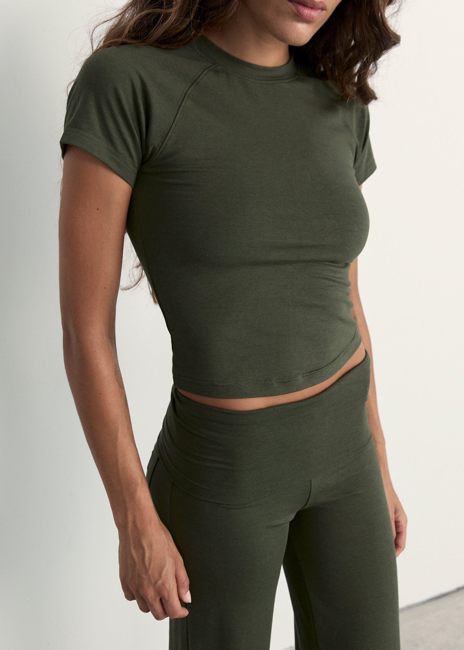 A woman wearing a fitted short-sleeve crop top and matching high-waisted leggings in olive green, shown from the shoulders to the upper thighs against a plain light background.