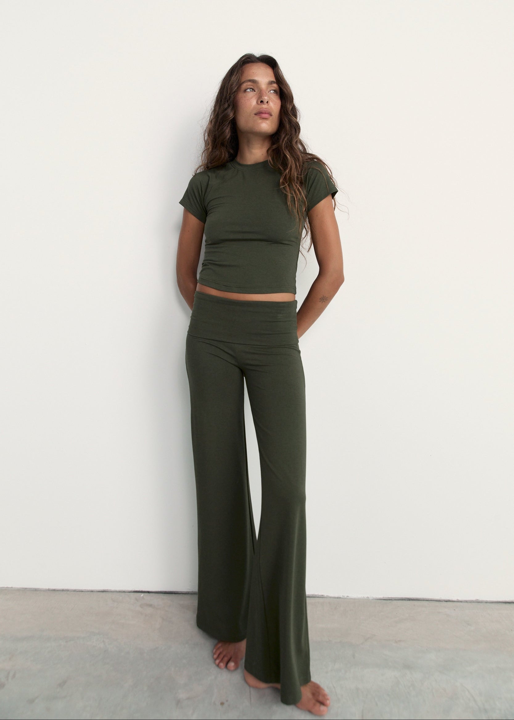 A woman stands against a plain white wall wearing an olive green two-piece lounge set consisting of a short-sleeve crop top and matching high-waisted flared pants.