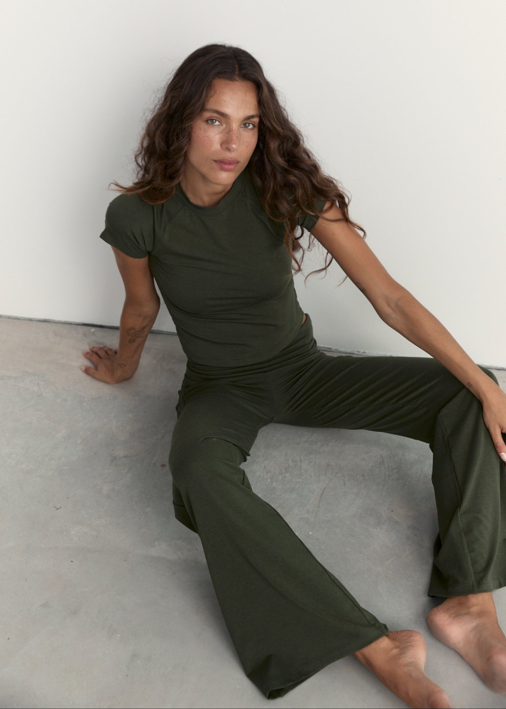A woman wearing an olive green short-sleeve top and matching wide-leg pants, seated barefoot against a plain white wall and light gray floor.