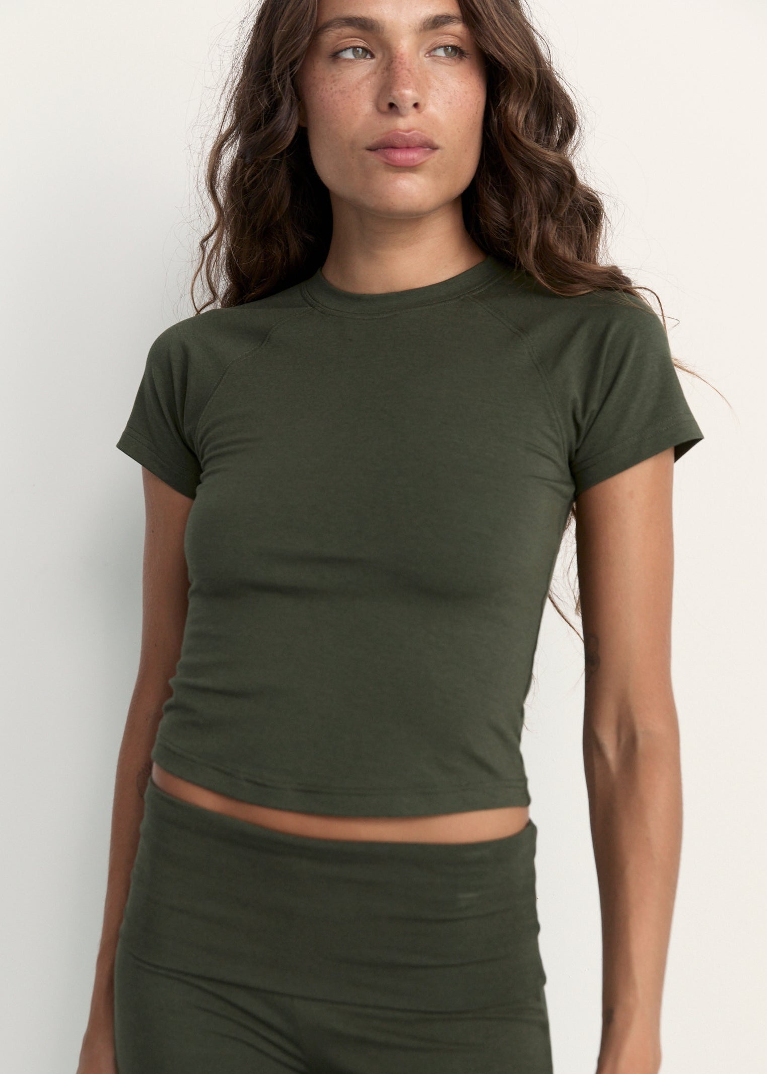 A woman wearing a fitted short-sleeve crop top in olive green, paired with matching green bottoms, photographed against a plain light background.