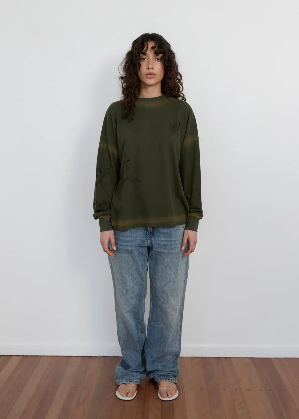 A model stands against a white wall wearing an olive green crewneck sweatshirt with subtle tonal embroidery, paired with wide-leg blue jeans and white sandals.