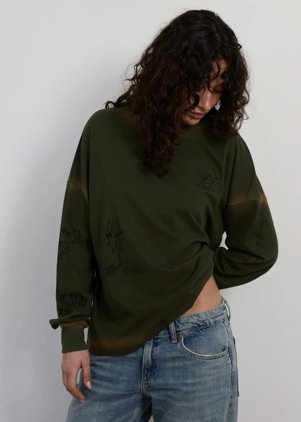 A woman wearing a dark olive green long-sleeve crop top with tonal embroidery and light wash blue jeans, standing against a plain light gray wall.