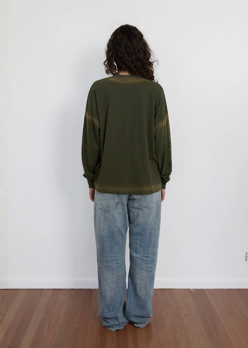 A person viewed from the back wearing an olive green long-sleeve top and light blue wide-leg jeans, standing on a wooden floor against a plain white wall.