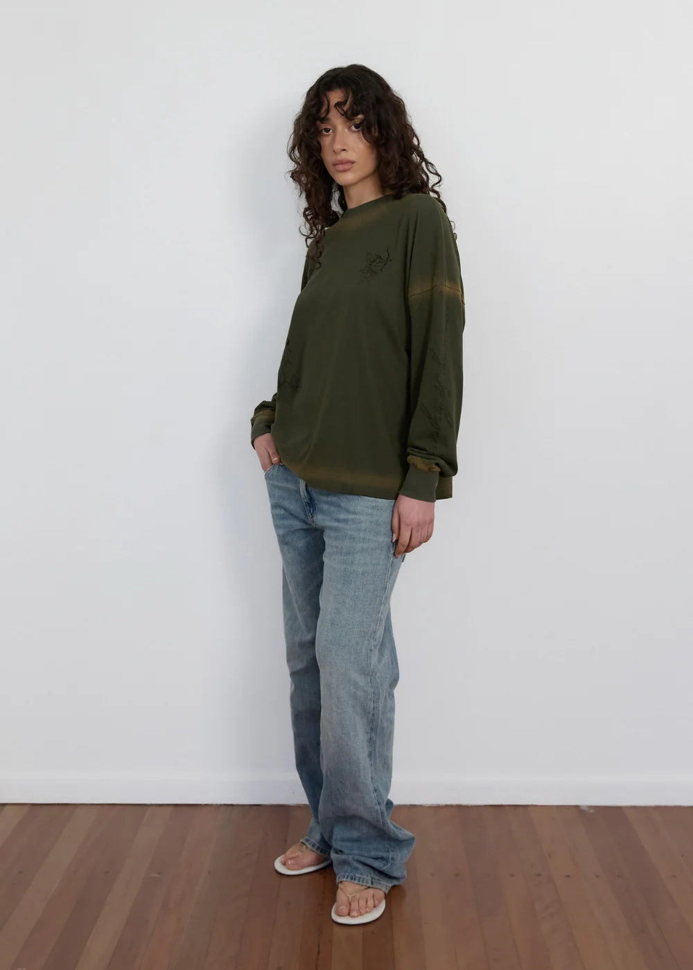 A model stands in a minimalist room with a white wall and wooden floor, wearing an olive green long-sleeve crewneck sweatshirt, light-wash blue jeans, and white sandals.