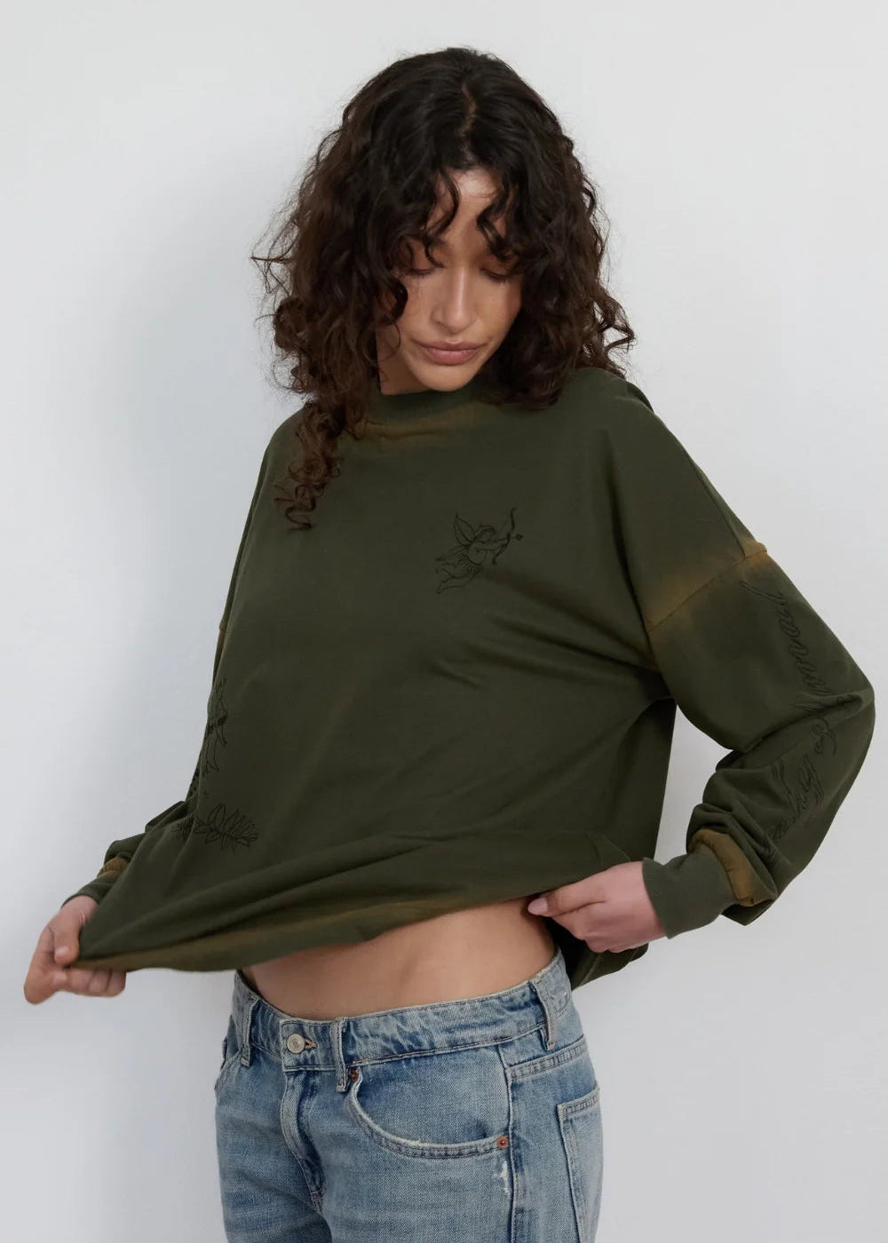 A woman wearing a dark olive green long-sleeve sweatshirt with distressed details and a faded brownish tint on the sleeves, paired with light blue jeans, against a plain light gray background.