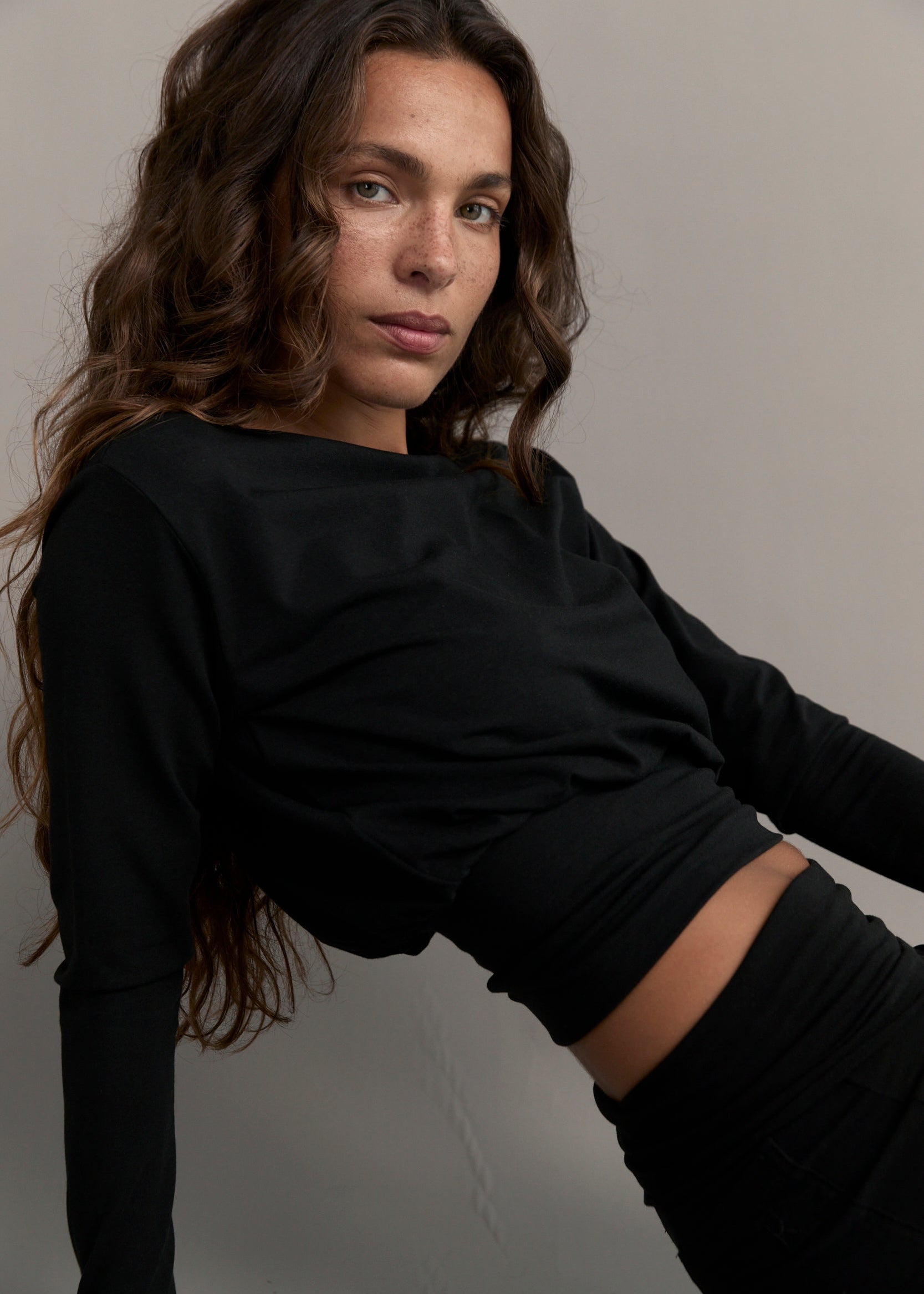 A woman with long wavy brown hair wearing a black long-sleeve crop top and matching black bottoms, posing against a neutral gray background.