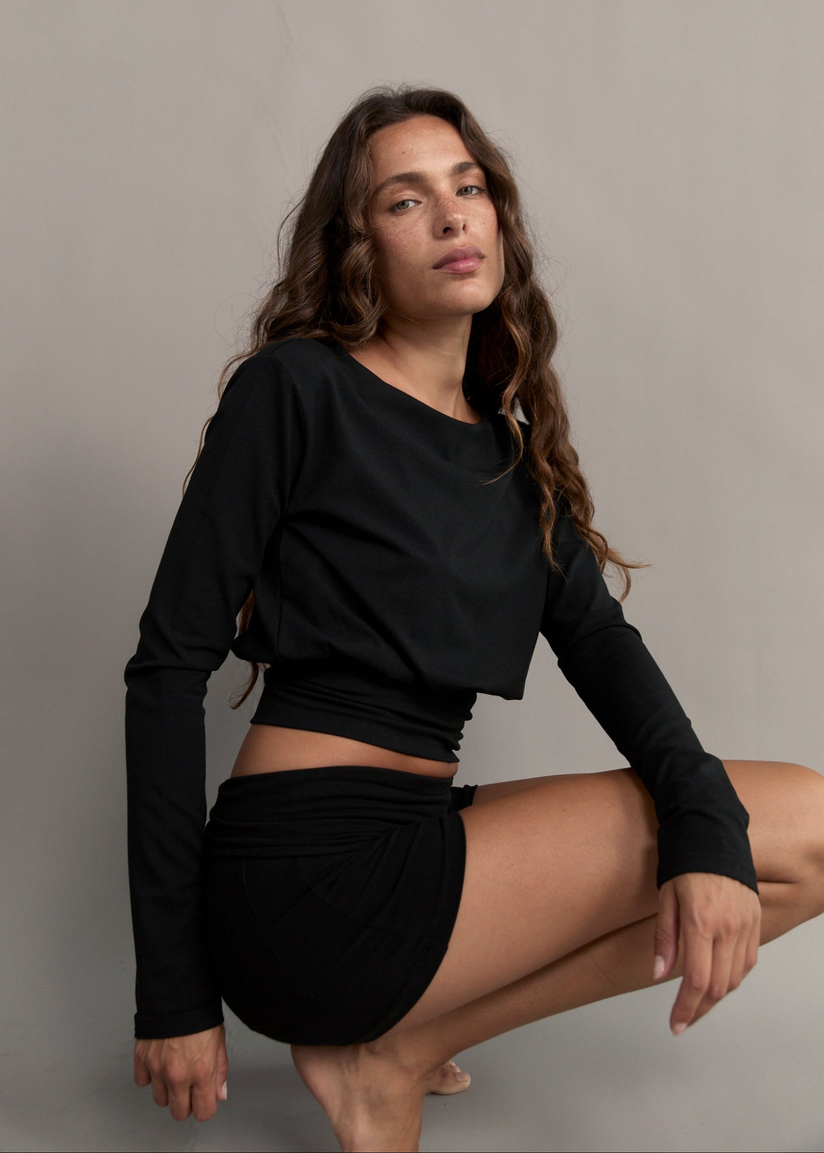 A woman crouching in a studio setting, wearing a black long-sleeve crop top and a matching black ruched mini skirt against a plain light gray background.