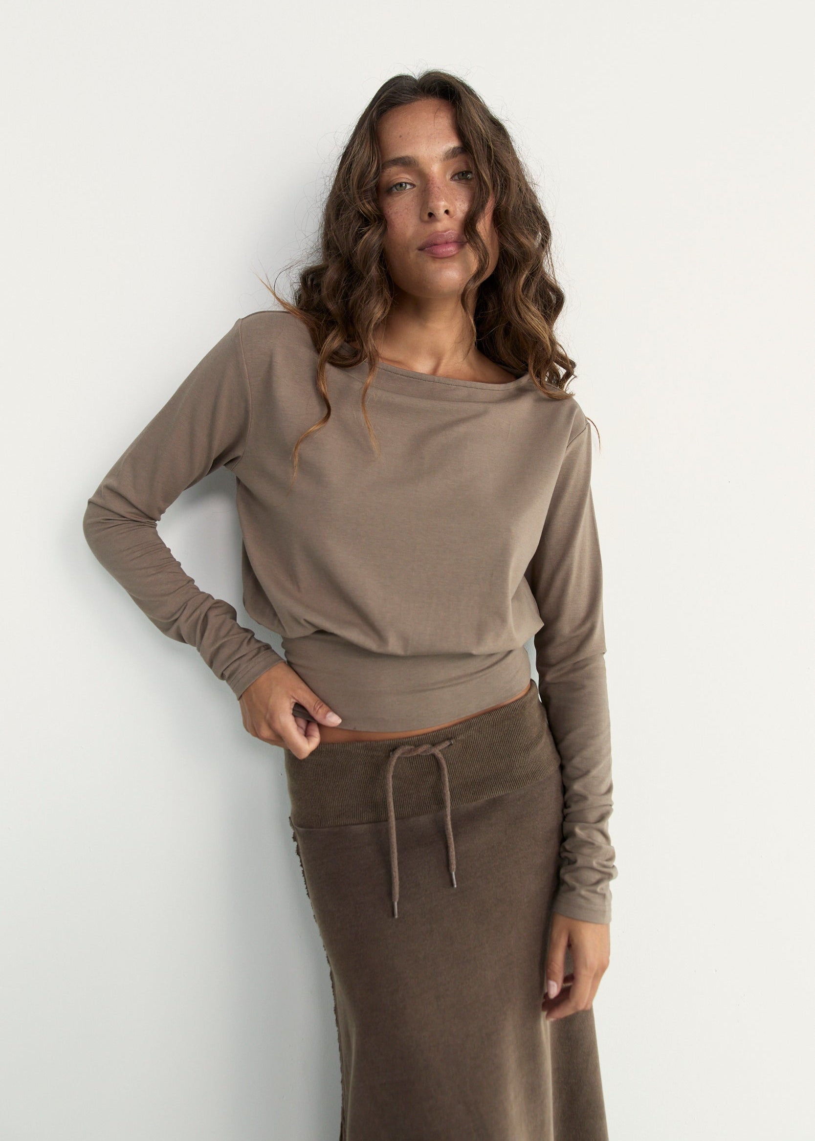 A woman modeling a taupe brown long-sleeve knit top and matching brown skirt against a plain white wall.